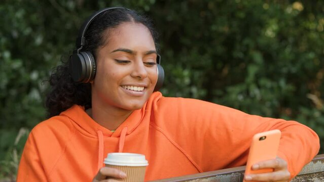 Beautiful mixed race African American girl teenager young woman listening to music on her smartphone or cell phone wearing wireless headphones and drinking take out coffee