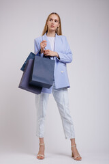 Fototapeta premium High fashion photo of a beautiful elegant young woman in a pretty blue jacket, blazer, denim jeans, shopping bags posing over white, soft gray background. Studio Shot. Slim Figure, Blonde