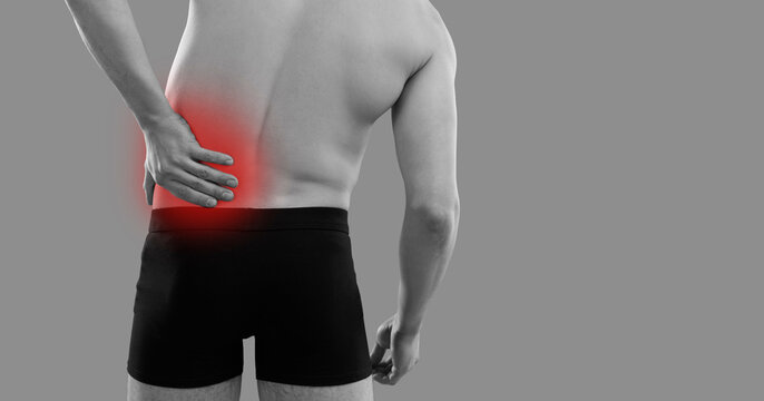 Man suffering from pain in his left side. Patient in underwear standing on grey background holding hand on his inflamed lower back with red highlight, backside rear view from behind. Backache concept