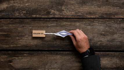 Hand of a businessman directing an origami made paper boat pulling a wooden peg with Follow us sign...