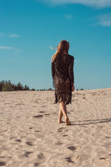 Girl with blonde hair in a black lace dress walking on the sand dunes of the desert with footprints in the sand, a young woman walking on golden sand on a bright summer day against a blue sky