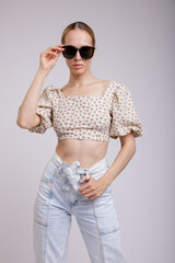 Naklejka premium High fashion photo of a beautiful elegant young woman in pretty blue denim jeans, beige top with floral pattern, stylish sunglasses, gathered hair on white, soft gray background. Slim figure. Blonde.