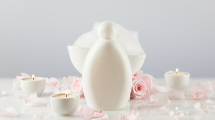 Spa relax composition with white cosmetic bottle, soap foam, candles and rose petals on white table. Wellness and skin care treatment. Mock up of bath beauty product. Concept of me time and self care
