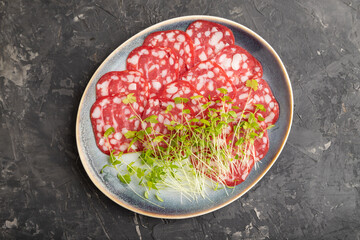 Slices of smoked cervelat salami sausage with spinach microgreen on black, top view, close up.