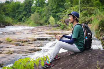 Asian young man, tall and slender, Wear your favorite blue hat, Sitting chilling on a large rock, enjoying the cool breeze,looked happy with  in front of him. Looking at hiking map from tablet in hand