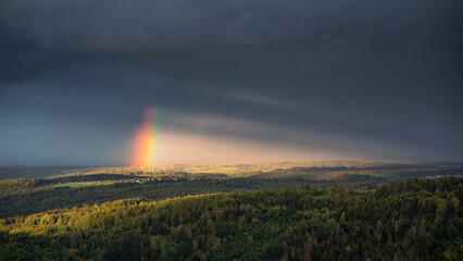 The sun shines through a gap in the cloudy sky and brings the end of the rainbow to light