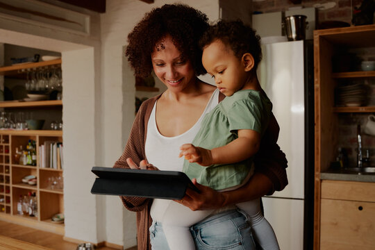 Biracial mother carrying daughter while looking down at digital tablet in kitchen at home