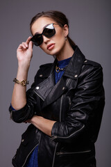 Fototapeta premium High fashion photo of a beautiful elegant young woman in a pretty leather jacket, blue dress, accessories, sunglasses, posing over gray background. Studio Shot. Portrait