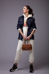 High fashion photo of a beautiful elegant young woman in a pretty white sports suit with hood, denim jeans jacket, sneakers, walking style, handbag posing over gray background. Studio Shot. Portrait.