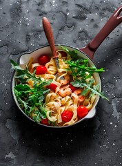 Fettuccine with shrimp, cherry tomatoes and arugula in a frying pan on a dark background, top view. Delicious Italian style lunch