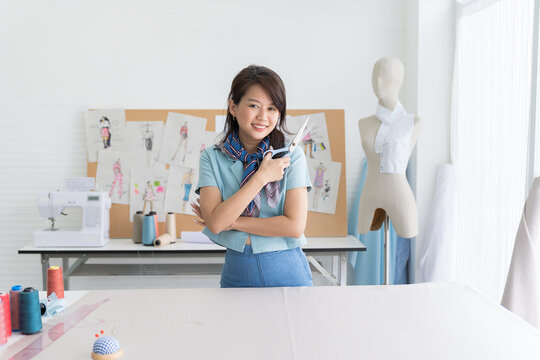 Portrait of happy asian female dressmaker holding scissors in the sewing workshop. Smiling woman seamstress working in tailor shop. Fashion, dressmaking or tailoring concept
