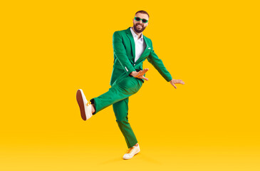 Fototapeta na wymiar Cheerful guy in a stylish party outfit dancing in the studio. Full length portrait of a happy man wearing a fashionable green suit and sunglasses dancing isolated on a bright yellow color background