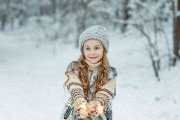 Fototapeta na wymiar Dreamy smiling young girl in knitted hat, mittens, sweater, vest with two burning christmas sparklers, bengal lights in her hands in winter forest with white snowy outdoor background