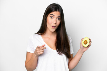 Young Brazilian woman holding an avocado isolated on white background surprised and pointing front