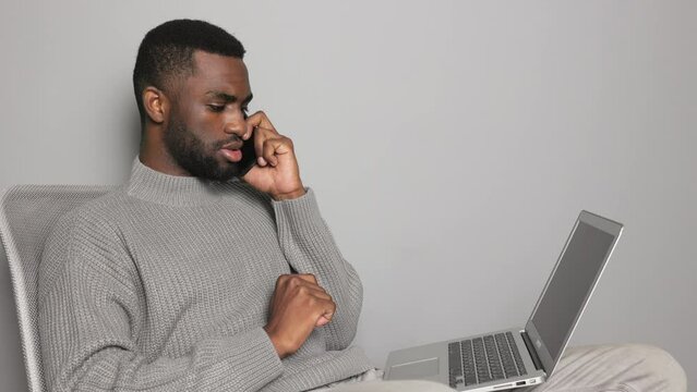African-American male freelancer sitting quarantined in a chair at home against a gray wall and holding his phone in his hand talking about work and looking at his laptop screen, work online