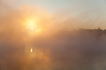 Sun shines through dense fog and reflects in still river, silhouettes of trees in fog, early morning. Ukraine.