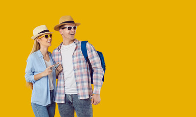 Positive young Caucasian man and woman in hats and sunglasses going on tour or mini travel during summer holidays, looking into distance with delight, taking pictures, stands on yellow background
