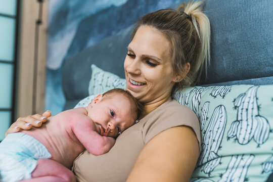 Infant baby in diaper slowly falling asleep while lying down on his mother's chest. Happy caucasian blond mother smiling at baby. Bedroom interior. High quality photo
