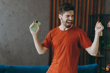 Young fun cool man in red t-shirt listen music in earphones use mobile cell phone dancing near blue sofa stay at home flat rest relax spend free spare time in living room indoor People lounge concept.