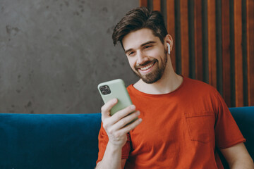Young man wears red t-shirt listen music in earphones use mobile cell phone sit on blue sofa stay at home flat rest relax spend free spare time in living room indoors grey wall. People lounge concept.