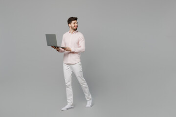 Full body young caucasian IT man 20s he wearing basic white shirt hold use work on laptop pc computer walk go look aside on workspace area isolated on plain grey background. People lifestyle concept.