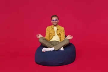 Ingelijste posters Zen man. Handsome calm focused guy sitting on bag chair in meditating lotus pose wearing blue shades, sunglasses and casual outfit isolated on red background. Happy calm freelancer enjoy free time © Svyatoslav Lypynskyy