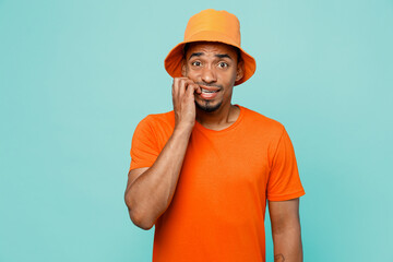 Young confused shocked sad man of African American ethnicity 20s wear orange t-shirt hat look camera biting nails fingers isolated on plain pastel light blue cyan background. People lifestyle concept.