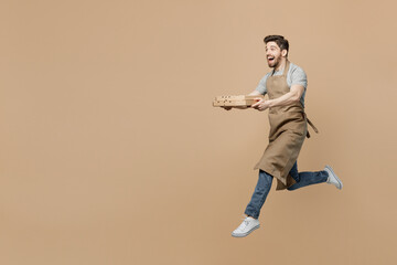 Fototapeta na wymiar Full size young man barista barman employee wear brown apron work in coffee shop jump run hold pizza in cardboard flatbox isolated on plain pastel light beige background Small business startup concept