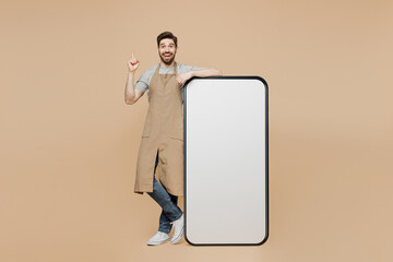 Full size young man barista barman employee in brown apron work in coffee shop big huge blank screen mobile cell phone point finger up isolated on plain beige background Small business startup concept