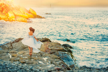 Romantic photo of a girl in a white dress on the ocean at sunset. View from the back.
