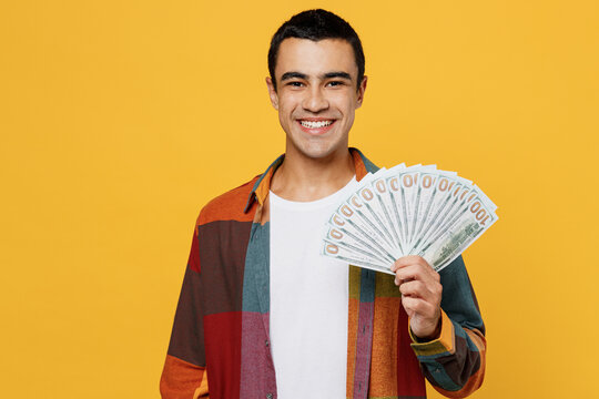 Young happy middle eastern man 20s he wear casual shirt white t-shirt hold in hand fan of cash money in dollar banknotes isolated on plain yellow background studio portrait People lifestyle concept.