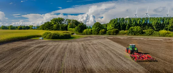 Wandcirkels tuinposter Aerial view of  farming tractor plowing on agriculture field after harvest with eolian wind farm in the background. © snapshotfreddy