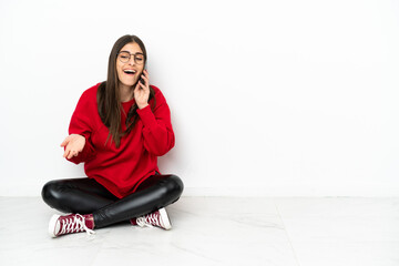 Young woman sitting on the floor isolated on white background keeping a conversation with the mobile phone with someone