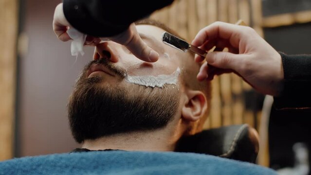 Professional stylist carefully shaves hipster beard with straight razor. Beard cut with old-fashioned sharp blade at barbershop. Handsome man getting his beard shaved in hair studio
