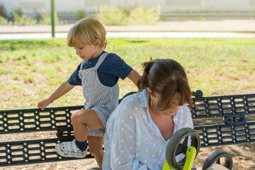 Little boy playing on a park bench while his grandmother repairs the tricycle so he can play.
