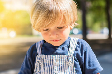 Portrait of an adorable 2 year old blond boy looking down, he is in the park on a sunny day with an out of focus background