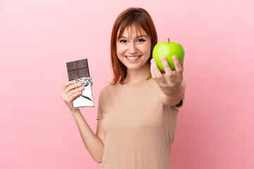 Redhead girl isolated on pink background taking a chocolate tablet in one hand and an apple in the other