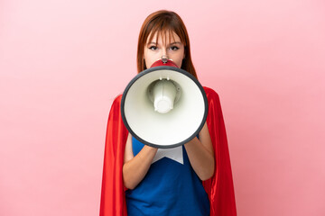 Super Hero redhead girl isolated on pink background shouting through a megaphone