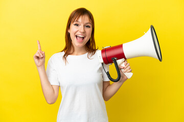 Redhead girl isolated on yellow background holding a megaphone and pointing up a great idea