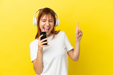 Redhead girl isolated on yellow background listening music with a mobile and singing