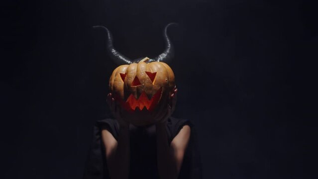 Horned demon lady wearing black dress holding scary carved pumpkin in front of her face, spooky jack o lantern, slowly revealing her beautiful face. Halloween costume and decoration concept