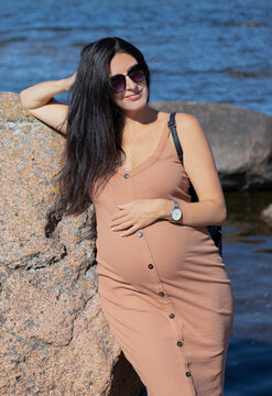 Heavily pregnant beautiful brunette woman with black sunglasses and beige tight dress leaning against a big boulder on the beach with a blue sea behind her on a sunny summer day