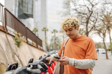 Handsome young caucasian guy using smartphone in bicycle parking lot. Blonde male hold hand on wheel outdoors. Concept gadgets, hobby, holidays.