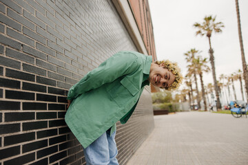 Funny young caucasian man in green shirt leans forward, stands outdoors. Curly blond looking at camera. Emotion concept, happy holiday time.