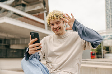 Fototapeta premium Affable young caucasian man waving at phone screen, speaking with friends, outdoor. Curly blonde smiling, wearing casual clothes atumun. Concept happy holidays.