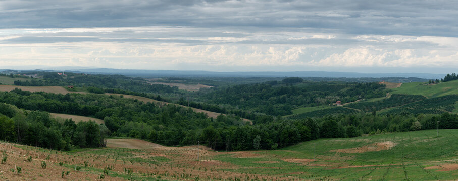 Panorama of hilly village with arable lands and forests, rural landscape during cloudy day in spring, bright cumulus clouds on horizon