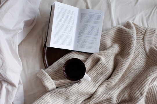 Open Paper Book With Cup Of Coffee On Wooden Tray And Knit Cloth Textile In Bed Close Up Top View. Cozy Home Atmosphere.