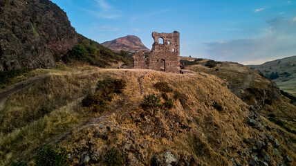 Ruin of St Anthony Chapel in summer evening with top of Arthur Seat at distance - drone photo