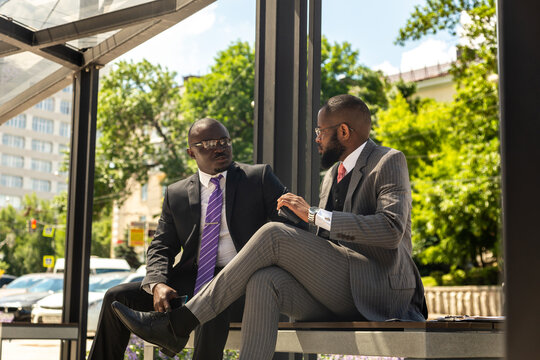 Two Two dark-skinned businessmen in suits communicate. Friends and business partners are sitting on a bench in the city against the backdrop of buildings