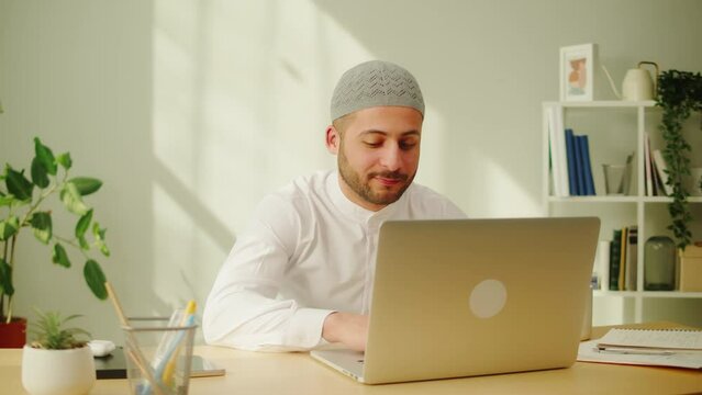 Middle eastern man using laptop. Home office. Male person texting at computer keyboard in living room. Wearing traditional Islamic clothes. Communicating with family and friends online.